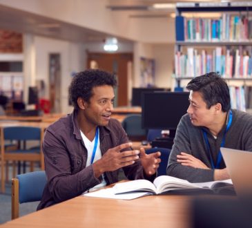 two male adult students talking at table in library