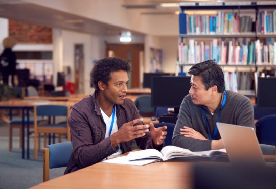 two male adult students talking at table in library