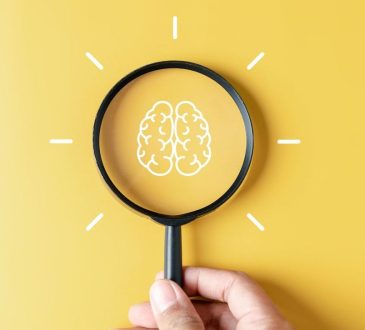Hand holding magnifying glass against yellow background with brain illustration in centre of lens