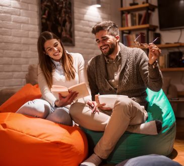 Happy couple smiling and reading book while sitting on lazy bags. Woman holding book. Living room interior.