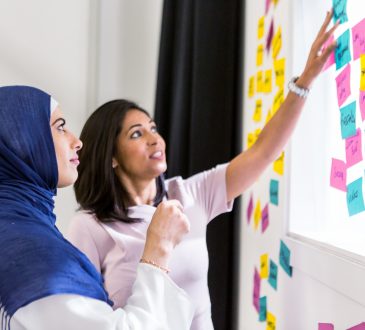 woman in hijab with woman not in hijab putting sticky notes on whiteboard
