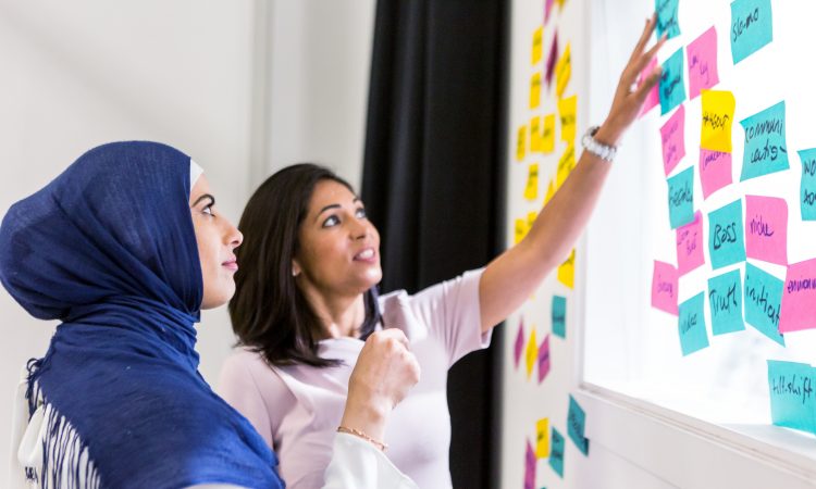 woman in hijab with woman not in hijab putting sticky notes on whiteboard