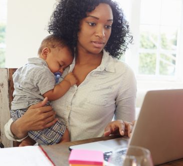 Mother With Baby Working In Office At Home Looking At Laptop