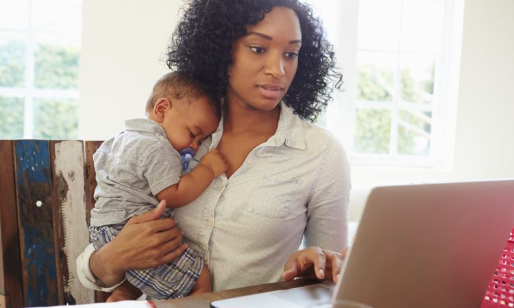 Mother With Baby Working In Office At Home Looking At Laptop
