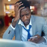 businessman looking stressed and resting his head on hand while typing message in smartphone sitting at counter in coffee shop,