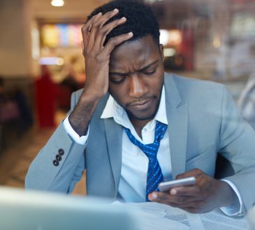 businessman looking stressed and resting his head on hand while typing message in smartphone sitting at counter in coffee shop,
