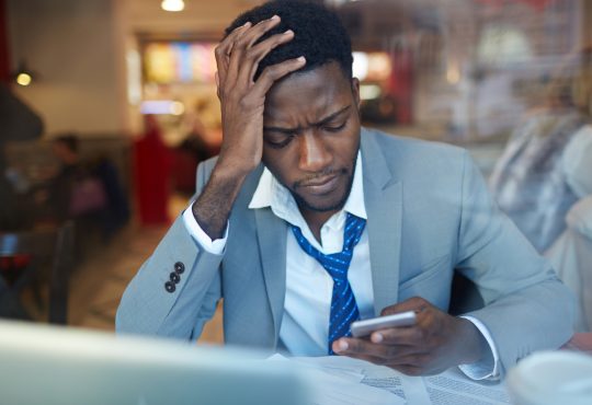 businessman looking stressed and resting his head on hand while typing message in smartphone sitting at counter in coffee shop,