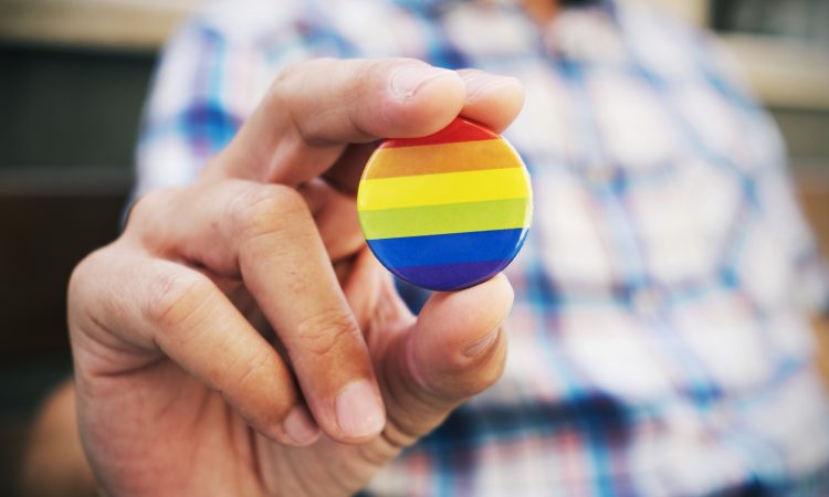 closeup of a young caucasian man outdoors holding a badge patterned with a rainbow flag