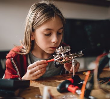 Girl wearing red hoodie looking circuit board throughout magnifying glass at home and building a robot