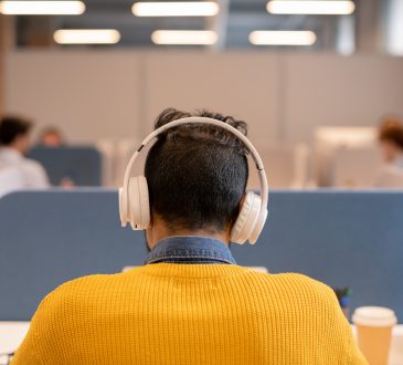 Rear view of busy brunette man in wireless headphones wearing bright sweater sitting at table in open space office a