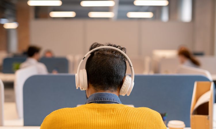 Rear view of busy brunette man in wireless headphones wearing bright sweater sitting at table in open space office a
