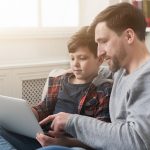 Happy father and son spending time together and surfing internet while sitting on sofa at home