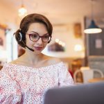 Portrait of young woman with headset and laptop indoors in cafe, working.