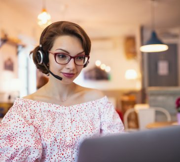 Portrait of young woman with headset and laptop indoors in cafe, working.