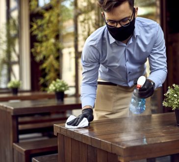 Waiter wearing protective face mask while disinfecting tables at outdoor cafe.