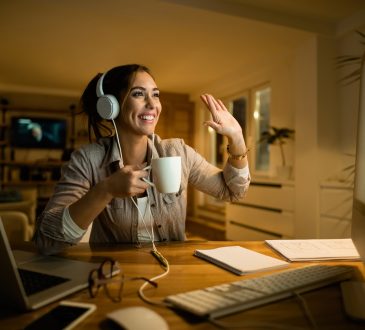 Happy woman drinking tea and waving to someone while having video call over desktop PC in the evening at home.