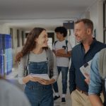Female high school student and male teacher walking in hallway talking amid other students.