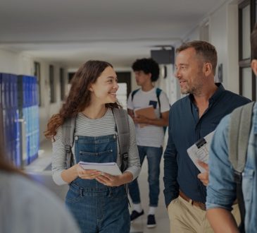 Female high school student and male teacher walking in hallway talking amid other students.