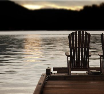 Adirondack chairs on a pier at sunset by the lake or ocean.