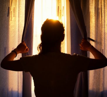 Silhouette of a young woman at the window opening curtains.