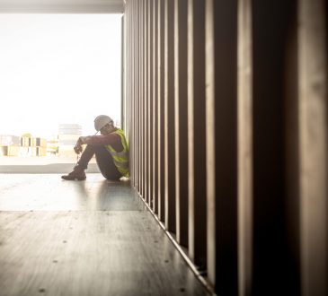 Man wearing hard hat and safety vest sitting with head hanging in container box at shipyard
