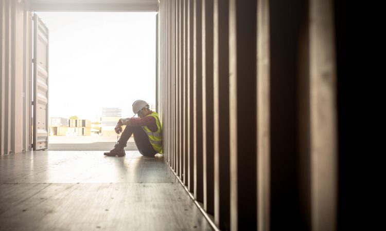 Man wearing hard hat and safety vest sitting with head hanging in container box at shipyard