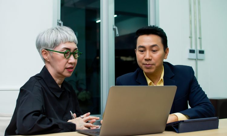 Older woman and younger male colleague talking over laptop in office