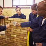 Teacher Helping Students Training To Be Builders Standing Next To Brick Wall