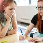 Two women meeting at table, with one writing while other looks on