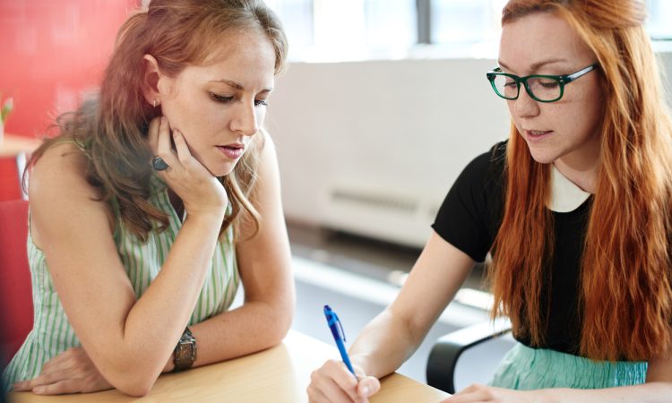 Two women meeting at table, with one writing while other looks on