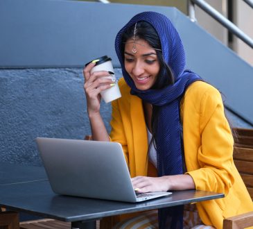 Young Indian businesswoman wearing yellow suit jacket smiling and working with a laptop at a coffee shop outdoors.