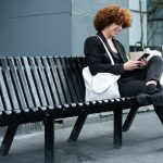 Non-binary person looking at phone on black bench outside of office