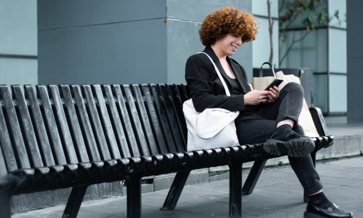 Non-binary person looking at phone on black bench outside of office