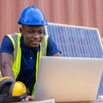 Engineer working on laptop outside in front of solar panel propped up against building