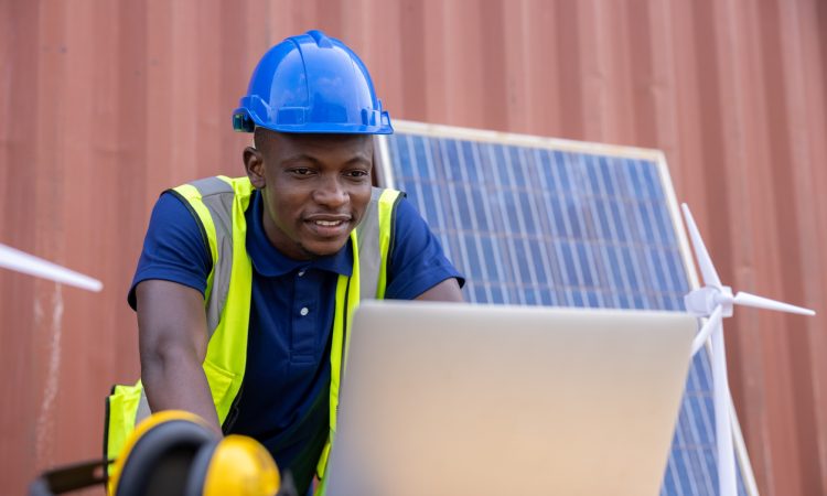 Engineer working on laptop outside in front of solar panel propped up against building