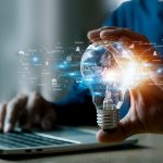 Person typing on laptop and holding glowing lightbulb in one hand with technology icons around it