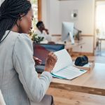Black woman with hair in braids reading documents at desk in office.