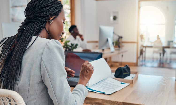 Black woman with hair in braids reading documents at desk in office.
