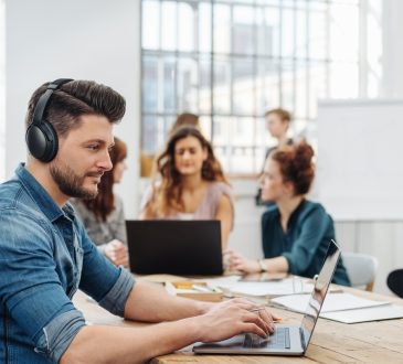Modern office with a dedicated young business team working on paperwork and laptops seated around a table with focus to a young man wearing headphones typing on a laptop