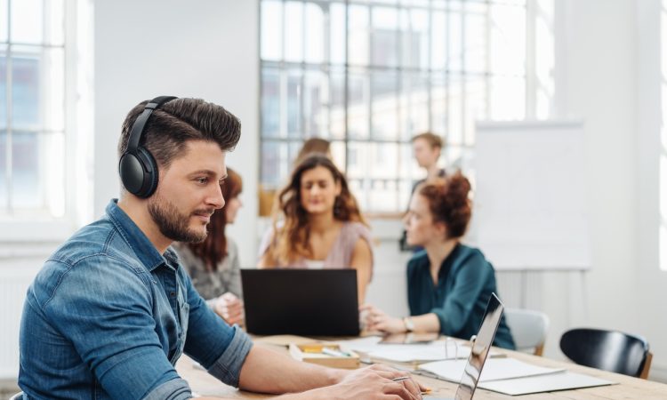 Modern office with a dedicated young business team working on paperwork and laptops seated around a table with focus to a young man wearing headphones typing on a laptop