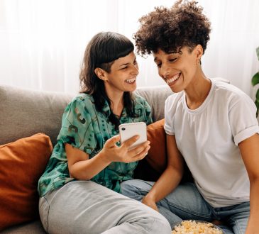 Lesbian couple laughing on couch together at home.
