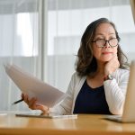 Older woman sitting at computer and examining paperwork