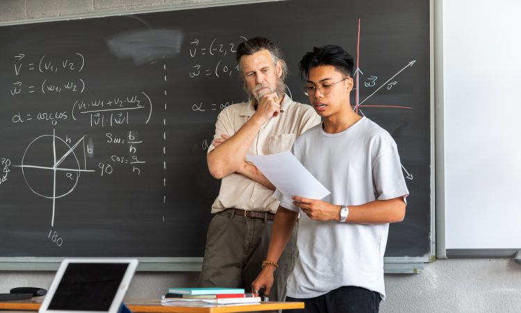 Student stands beside teacher at front of classroom looking at notes.