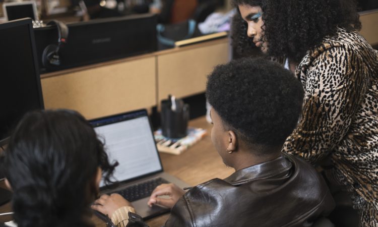 Three co-workers of varying genders working on a laptop, seen from back. The person on the right, whose face is most visible, is wearing teal eyeshadow. They are Black and have shoulder-length, curly brown hair. They are wearing a leopard-print blazer.