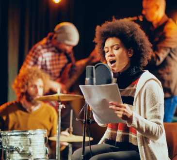 Woman with brown afro hold paper and sings, with band laying in background