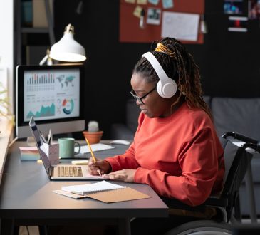 A Black woman with hair in braids in high ponytail, wearing white over-ear headphones and red long-sleeved shirt, sits in a wheelchair at a desk, taking notes in front of a laptop.
