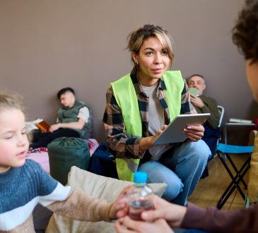 Young female volunteer wearing yellow vest squats in front of woman with son in community centre and enters their information on a tablet.