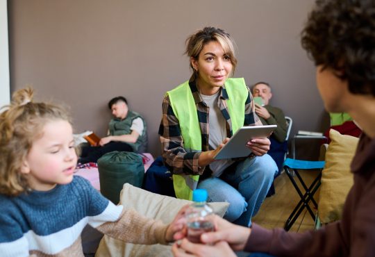 Young female volunteer wearing yellow vest squats in front of woman with son in community centre and enters their information on a tablet.