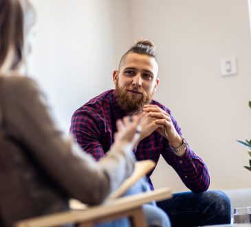 Man and woman sitting in chairs facing each other and talking. Woman is pictured from behind, with brown blazer and blonde hair. Man is wearing blue and red plaid shirt. He has a bushy brown beard, hair shaved on the sides and a top knot.