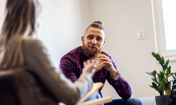 Man and woman sitting in chairs facing each other and talking. Woman is pictured from behind, with brown blazer and blonde hair. Man is wearing blue and red plaid shirt. He has a bushy brown beard, hair shaved on the sides and a top knot.